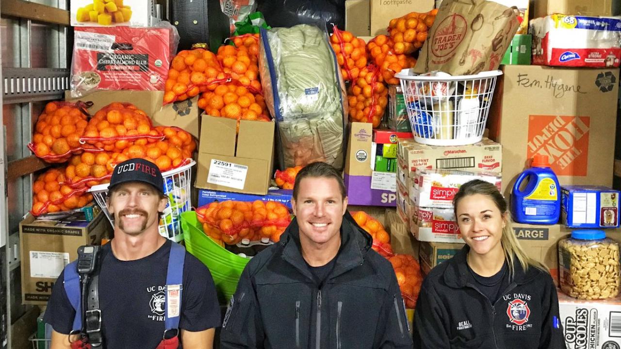 Three firefighters sit in front of a wall of donated items.