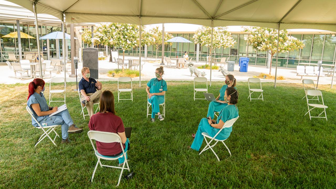 Dean in polo shirt chats with five woman in scrubs under a tent.