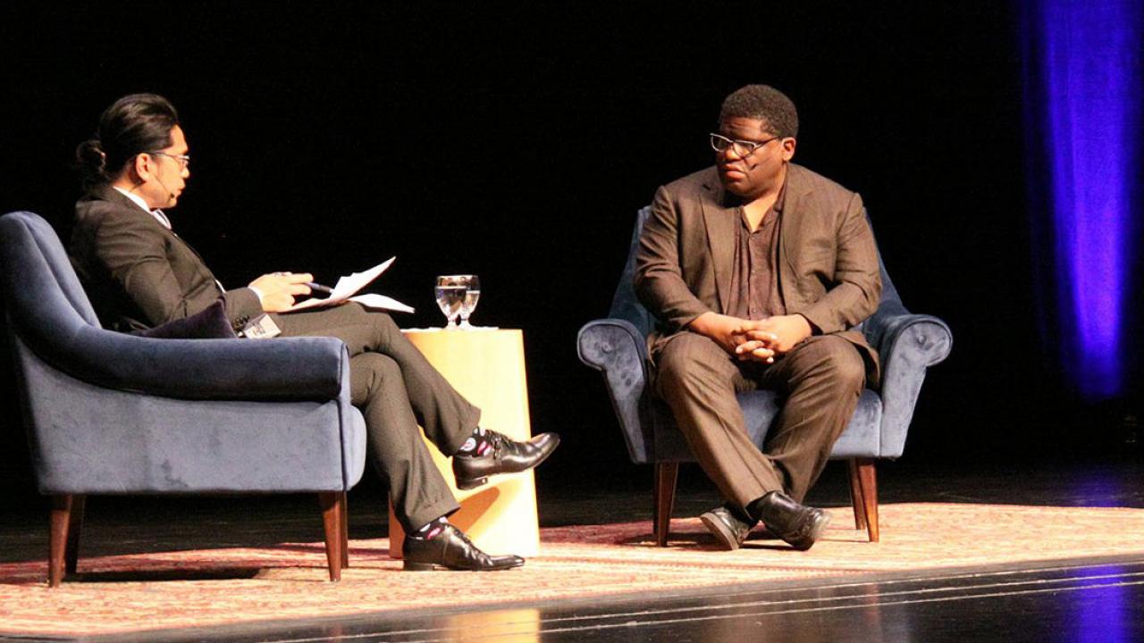 Two men, in chairs, on stage, in conversation.