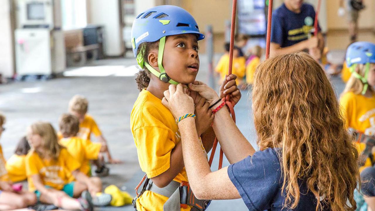 Camp leader puts helmet on girl at rock climbing wall.