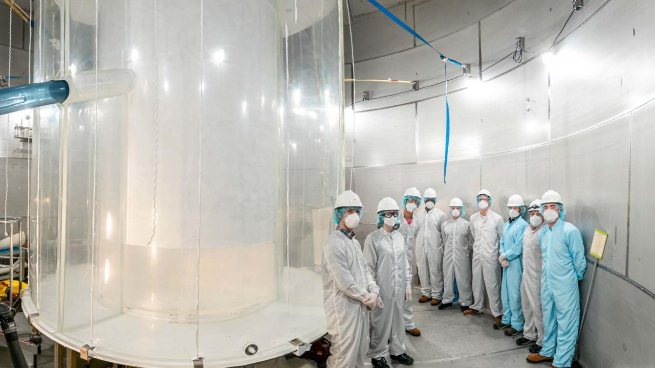 A large translucent tank fills the left side of the photo. A group of people in coveralls, helmets and facemasks stands against the wall. 