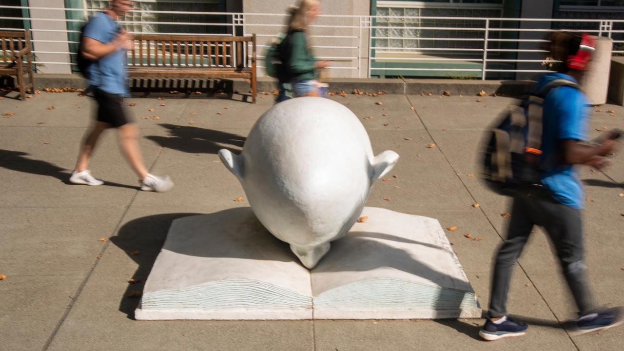 Bookhead Egghead shot in front of library at UC Davis with students, in blur, walking by
