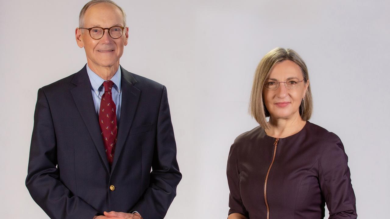 On the left, a white male with grey hair and glasses wearing a dark suit and red tie. On the right is a white woman with blonde hair and glasses wearing a deep purple blouse. 