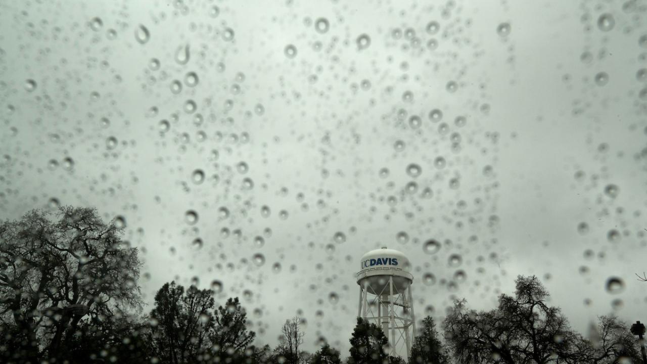 landscape of UC Davis water tower as seen through rain-splattered window on a gray day with above a silhouette of trees.
