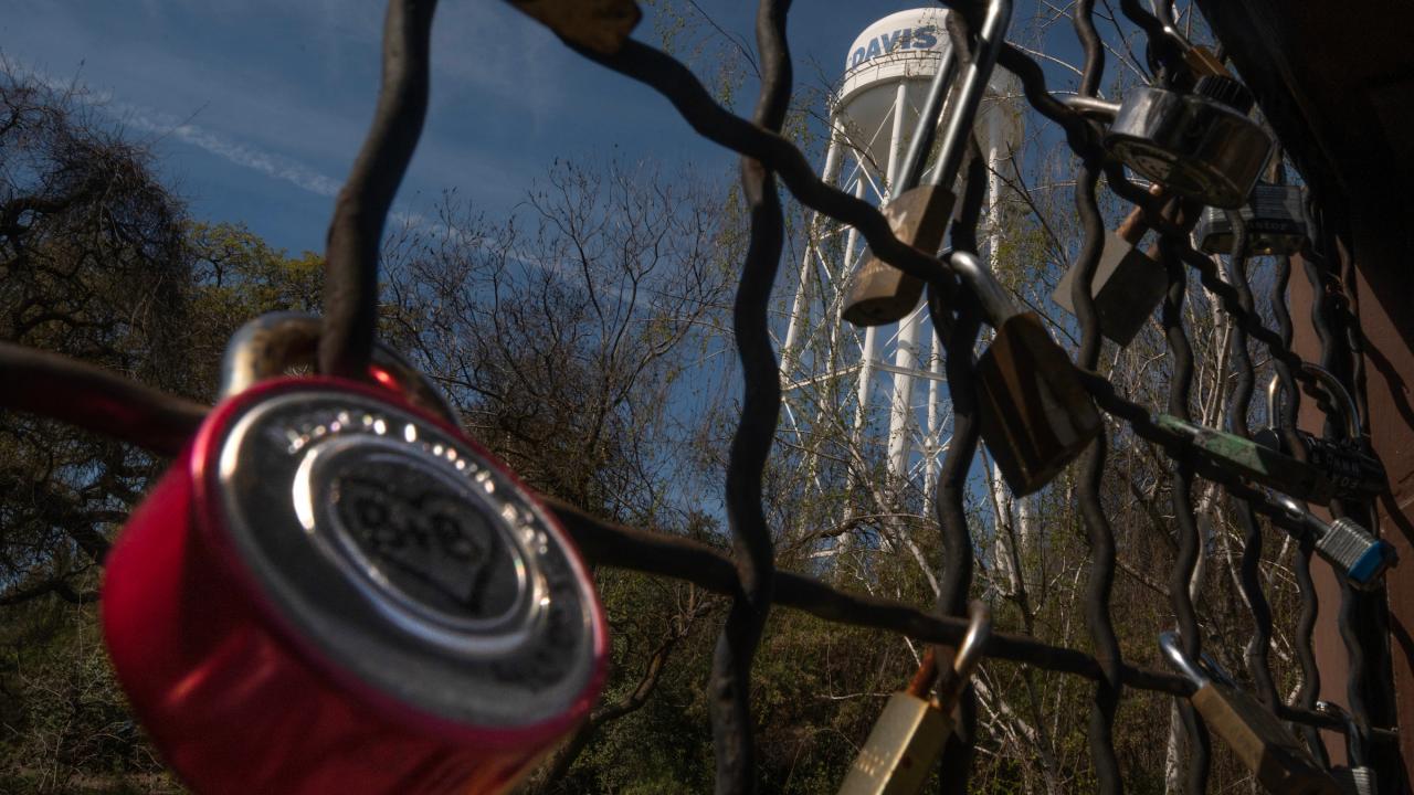 A close-up of a red padlock on a fence, with a water tower visible in the background.