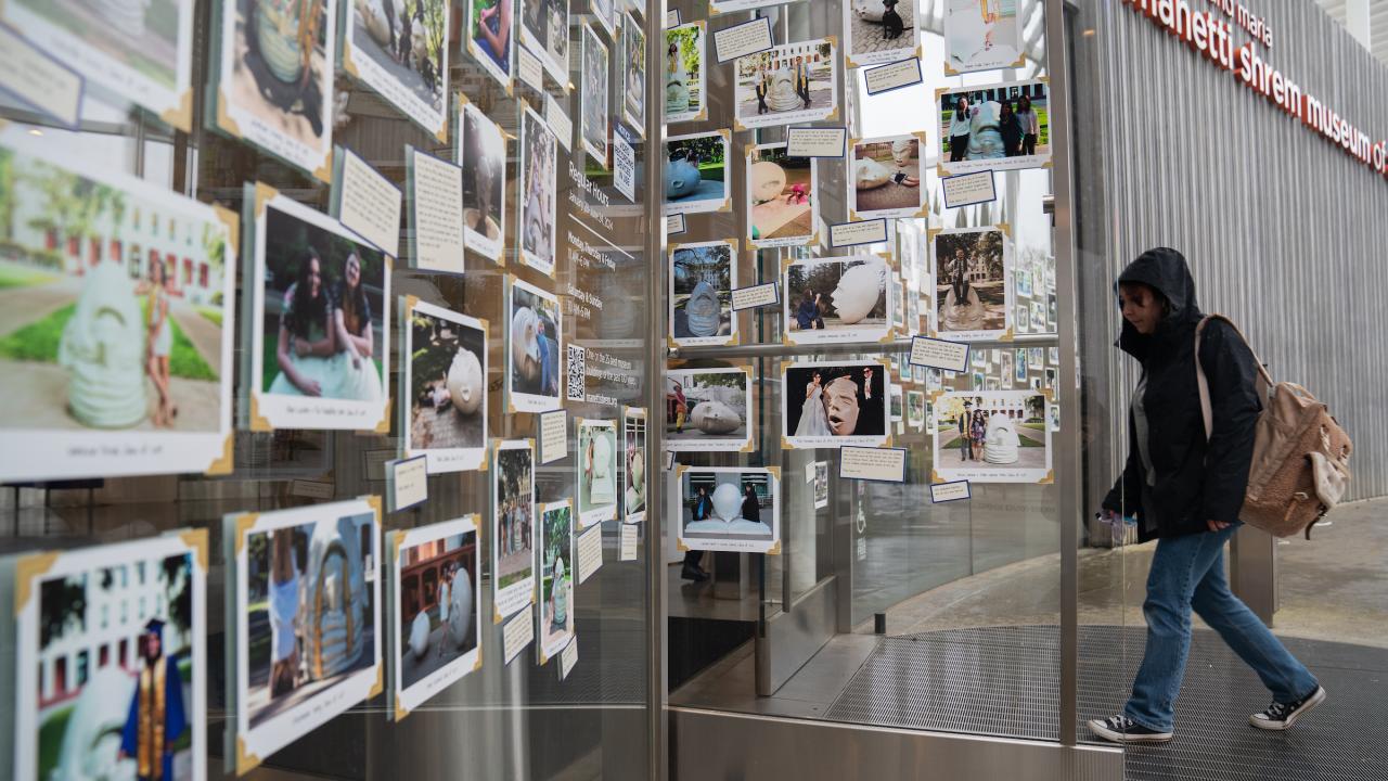 On a rainy day, a student in a black parka opens a glass door and enters a museum lobby. The glass door and its nearby wall are covered with photographs of UC Davis students standing next to the Egghead sculptures created by Robert Arneson.
