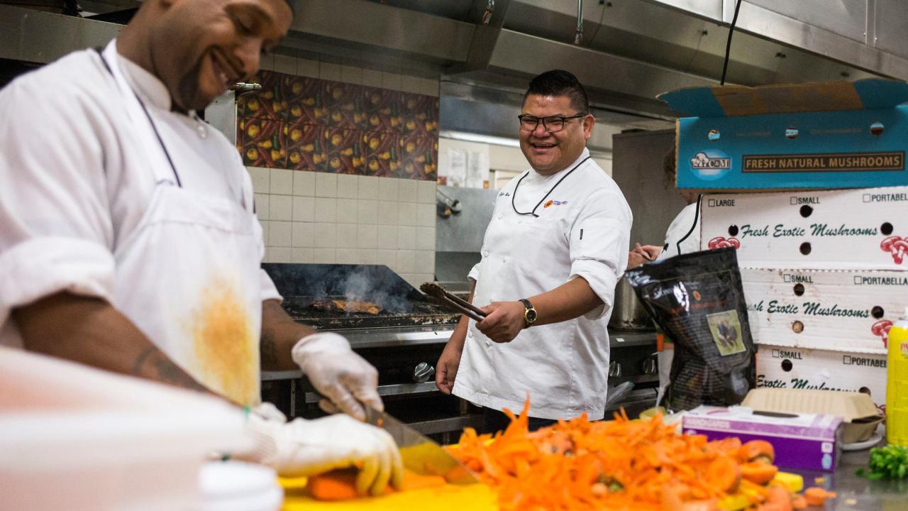 Two chefs are seen laughing while cooking inside an industrial kitchen. Chef Freddie Bitsoie is on the far right, facing the camera, in front of the grill.