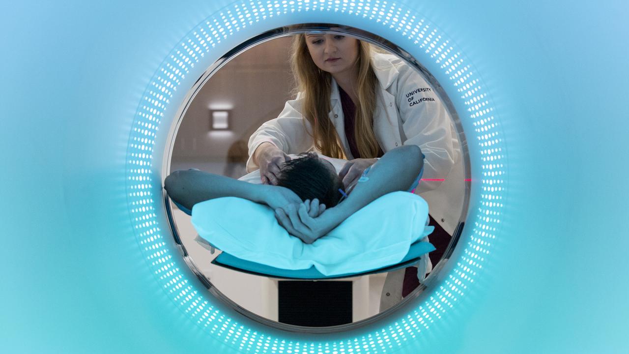 In a circular opening, a long-haired woman in a white lab coat bends over a person lying down with the top of their head towards the camera. Blue color fills the rest of the field outside the circle. 