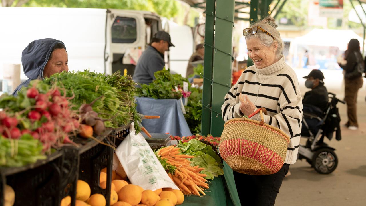 Woman holding woven basket chats with vendor at farmers market stall of carrots and greens