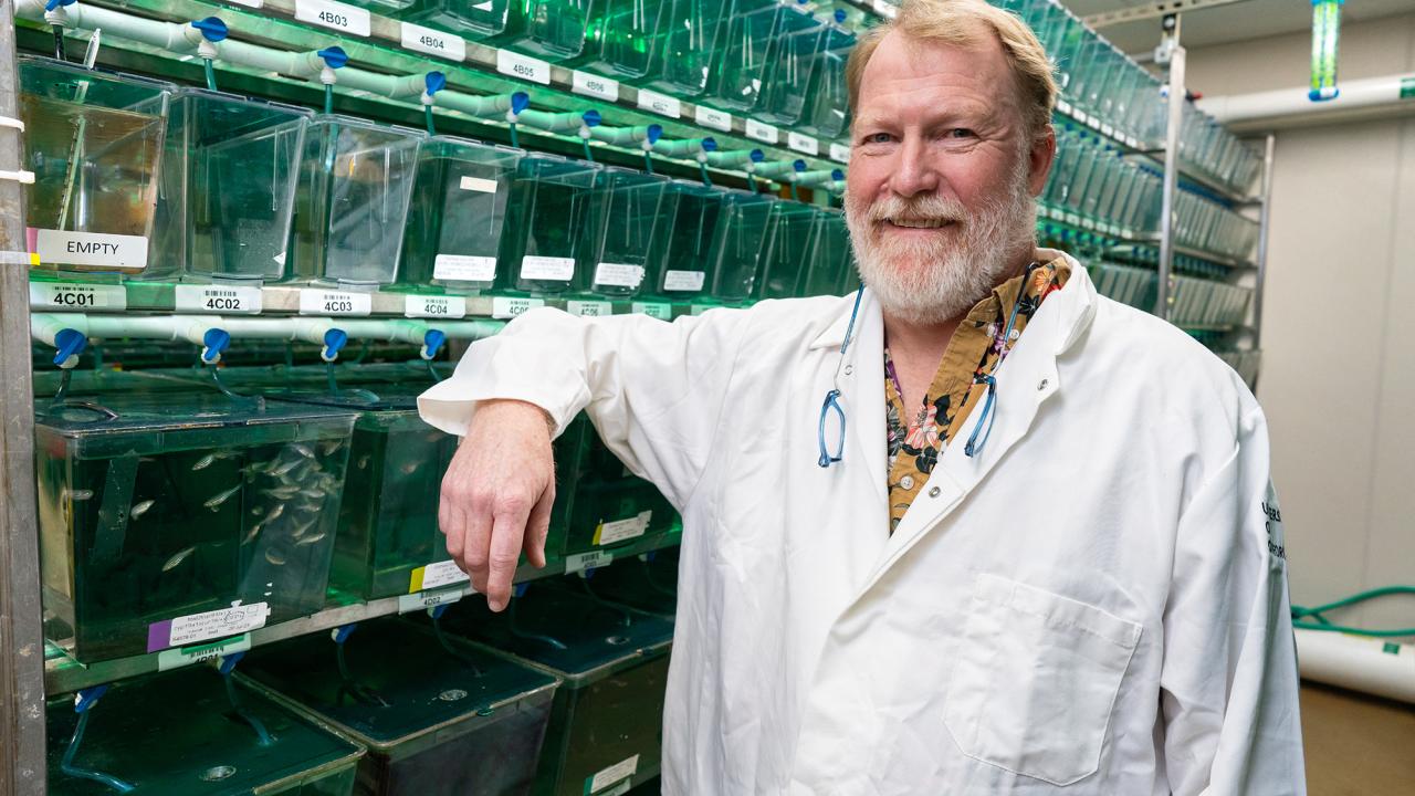 White man with ginger hair and beard wearing a white labcoat standing in front of rows of fish tanks. 