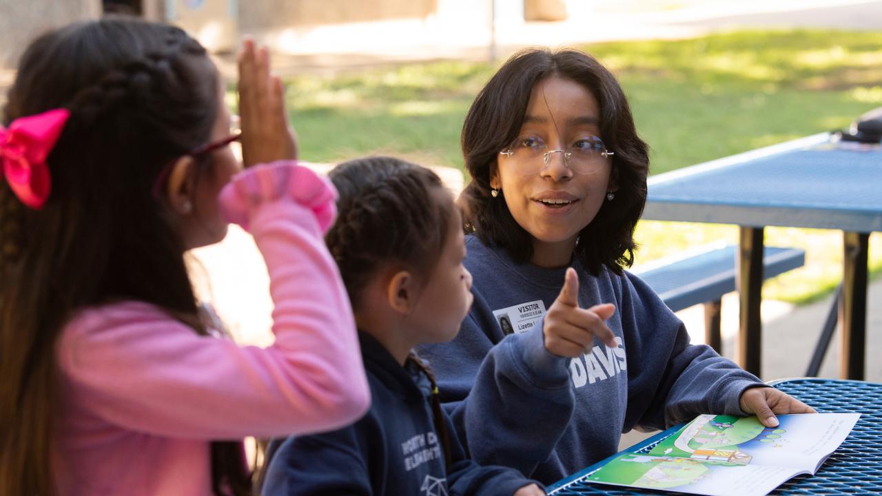 A female university student points at a child raising her hand during a reading lesson at an outdoor picnic table.