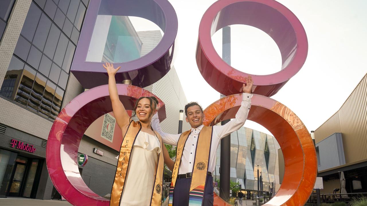 Two UC Davis students in graduation stoles in front of the colorful DOCO sign in downtown Sacramento.