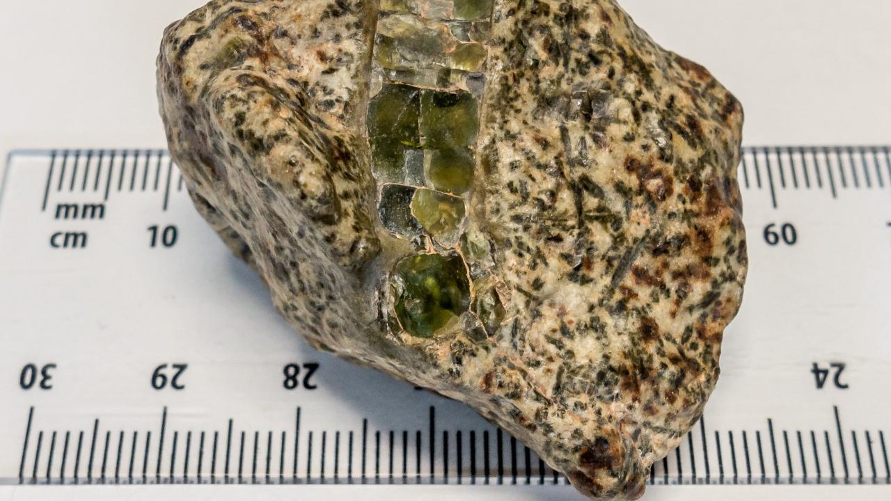 Roughly triangular pale off-white rock with brown and black flecks and a band green crystal running vertically across it. A ruler shows that it is about 50 millimeters across. 