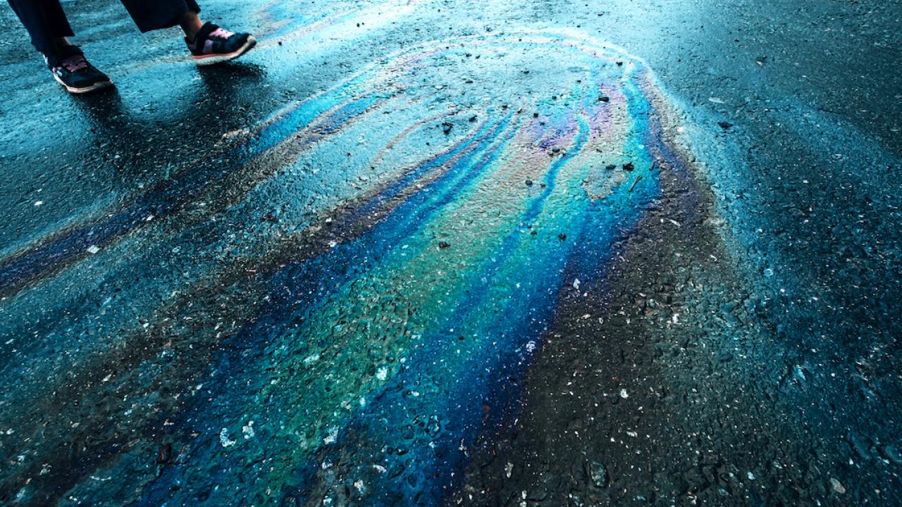 a rainbow sheen of oil on wet pavement and the legs of someone standing near it
