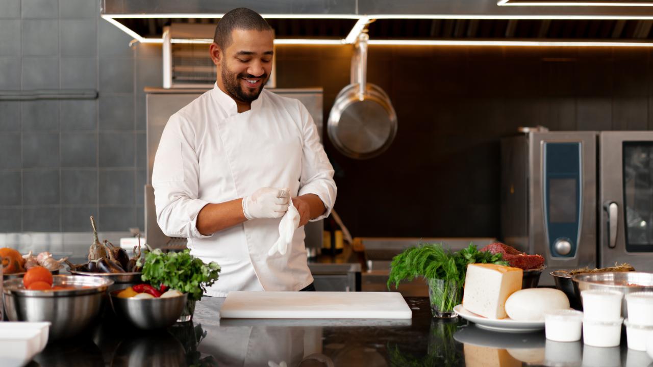 A male chef in a white coat puts white rubber gloves on his hands. He is standing in a kitchen behind a black countertop that has a white chopping board, raw meat and vegetables on it.