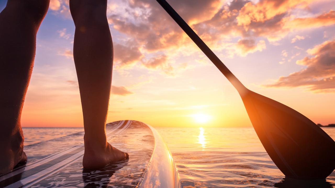 Legs shown of person on paddleboard in water toward sun