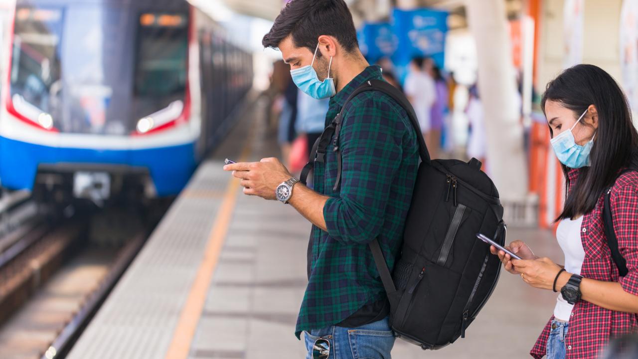 A man and woman wearing masks and looking at their phones wait to board an approaching train