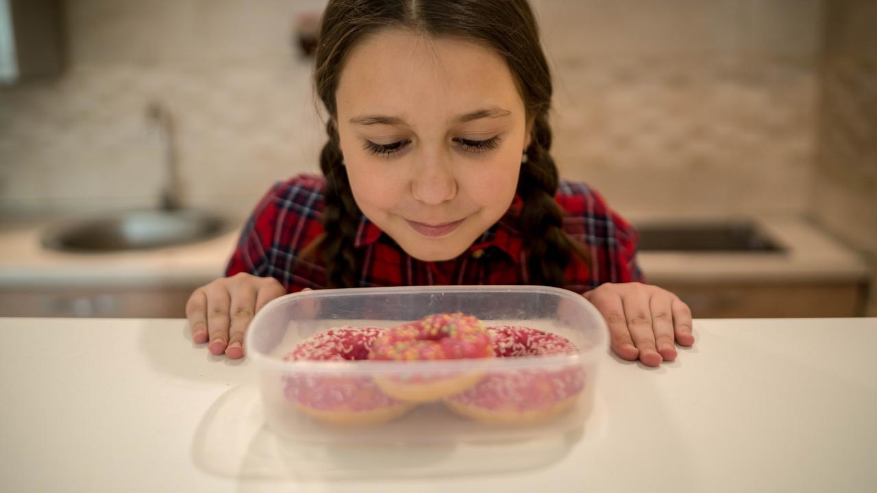 A child stands in a kitchen facing the camera, looking down into a tub of three pink glazed donuts.