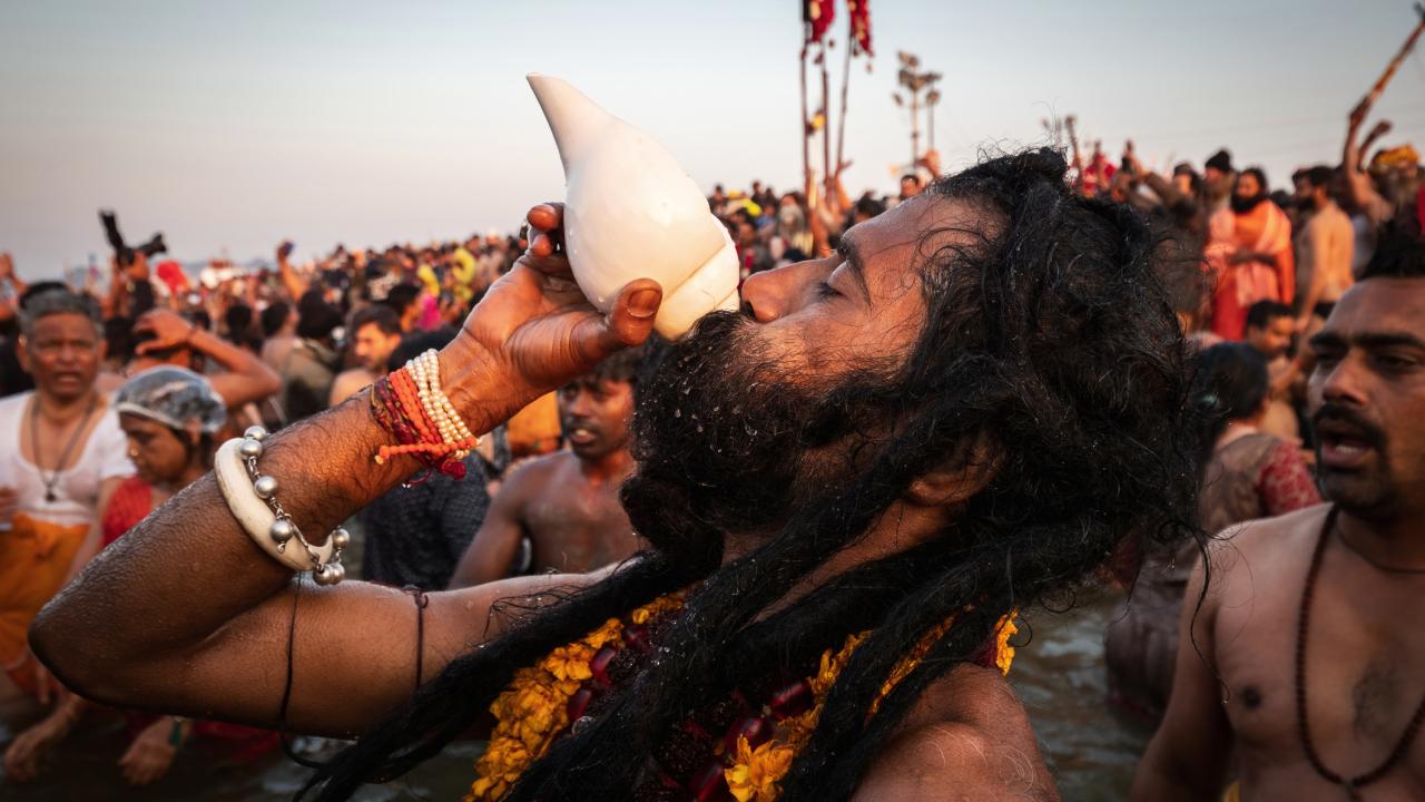 Man participates in festival in India, blowing into conch shell, while others look on 