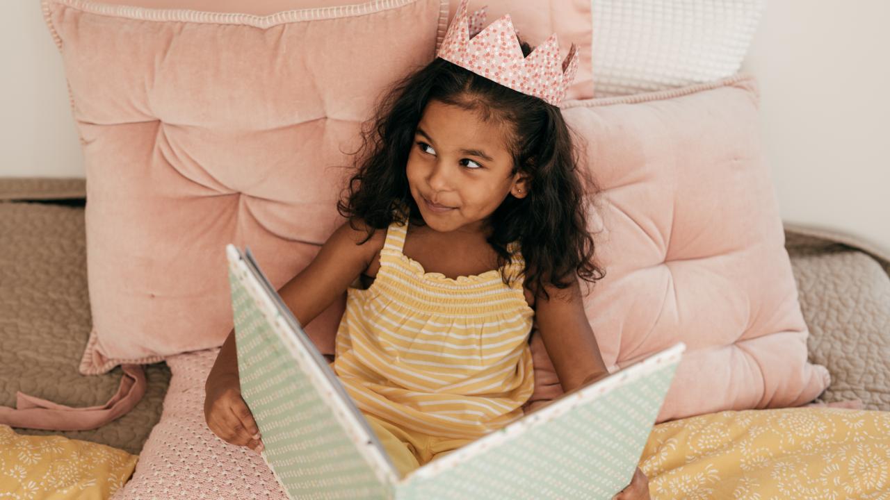 Young dark-complected girl with crown on head in pink-decorated bed looking at oversized book