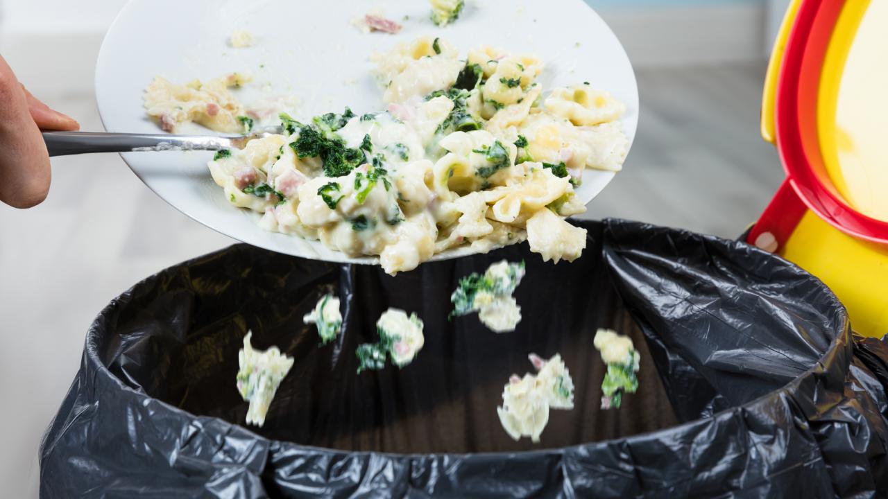 person's hand scraping plate of pasta and vegetables into trash can