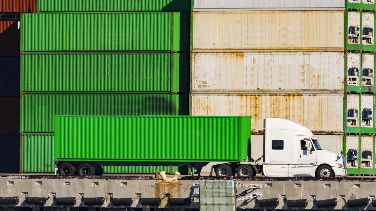 bright green heavy-duty truck sits parallel to stack of cargo containers at shipping port