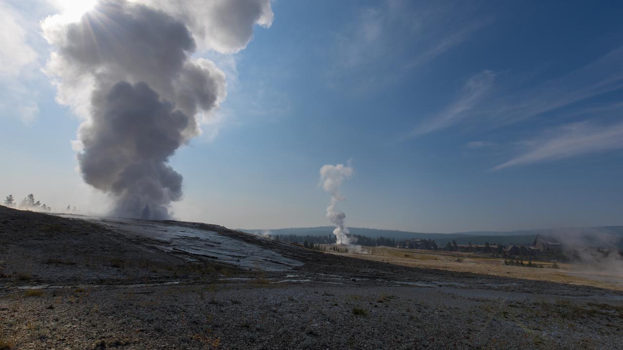 On the left, a large cloud of vapor rising from the ground with another geyser erupting in the background, against a blue sky. 
