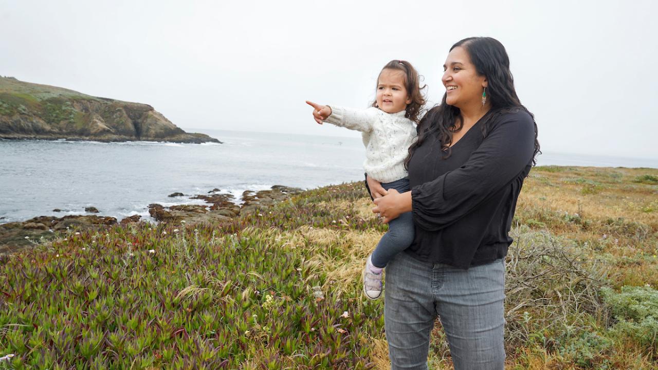 Woman in black shirt holds toddler daughter who is pointing at something. Ocean in background.