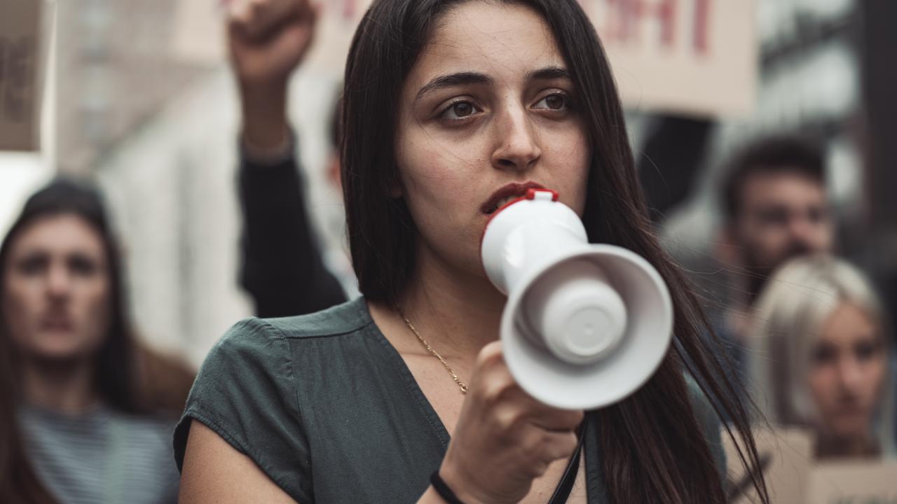 An Iranian female protestor holds a megaphone to her mouth. Behind her are blurred out protestors holding signs.