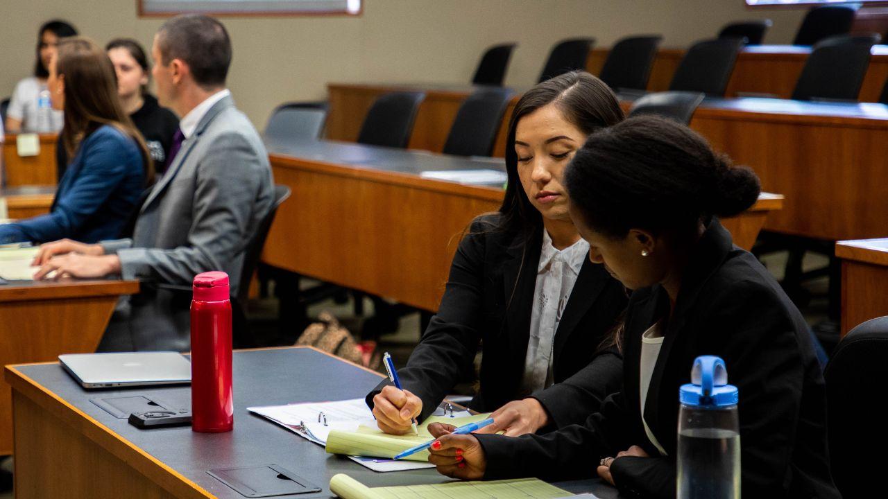 Two students in business attire sit at a desk. They deliberate together over a yellow legal pad.