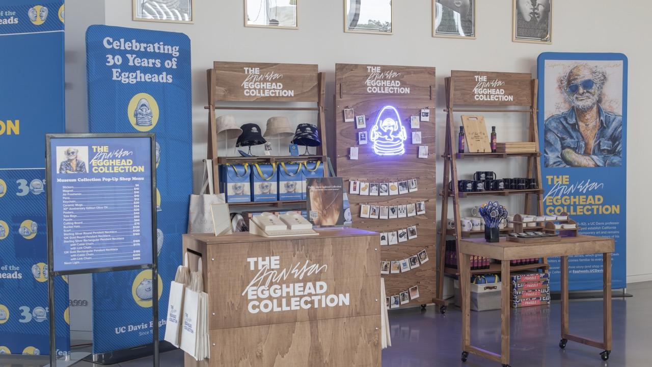 The Arneson Egghead Collection Pop-Up Shop at the Manetti Shrem Museum features three vertical banners featuring "Year of the Egghead" logo and image of Robert Arneson's self portrait, California Artist. In the center are three vertical display cases featuring items from the Collection, including bucket hats, pendant necklaces, black coffee mugs and even a blue neon sign in the shape of Arneson's sculpture "Eye on Mrak (Fatal Laff)"