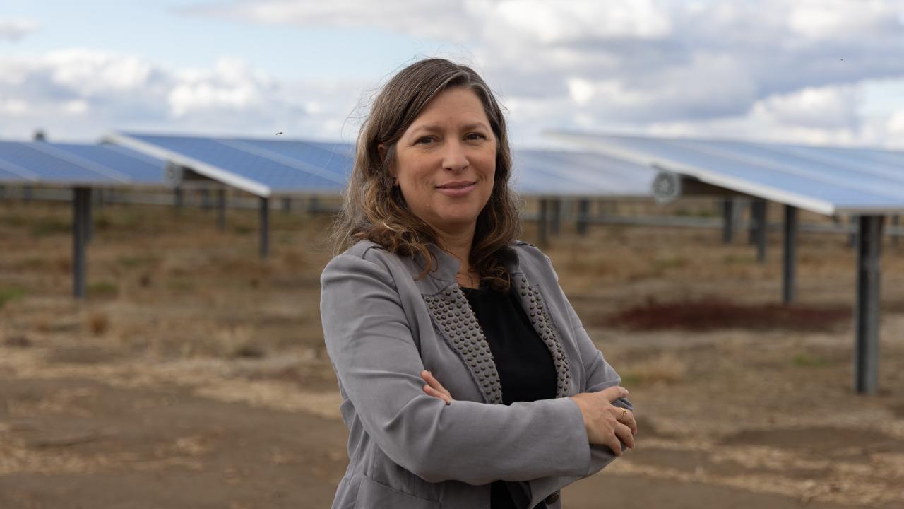 Professor Marina Leite in gray blazer, arms crossed, stands before rows of solar panels under cloudy sky