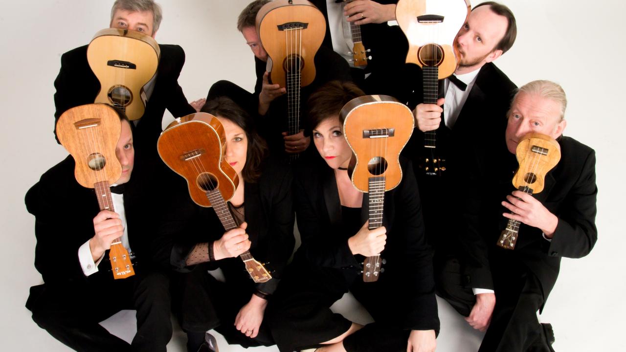 Eight orchestra members sit against a white background and hold a ukulele next to their face.