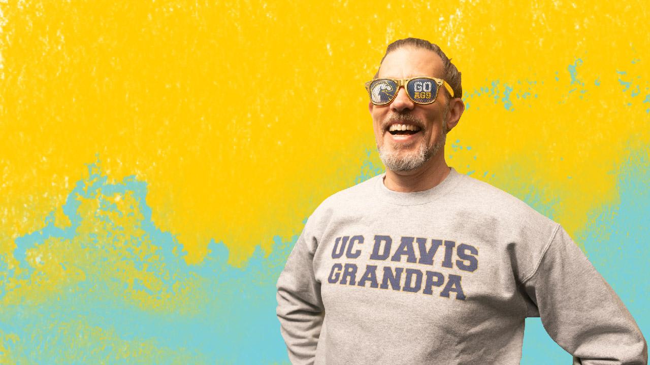 A person posing with an alumni sweatshirt in front of a yellow and green watercolor background