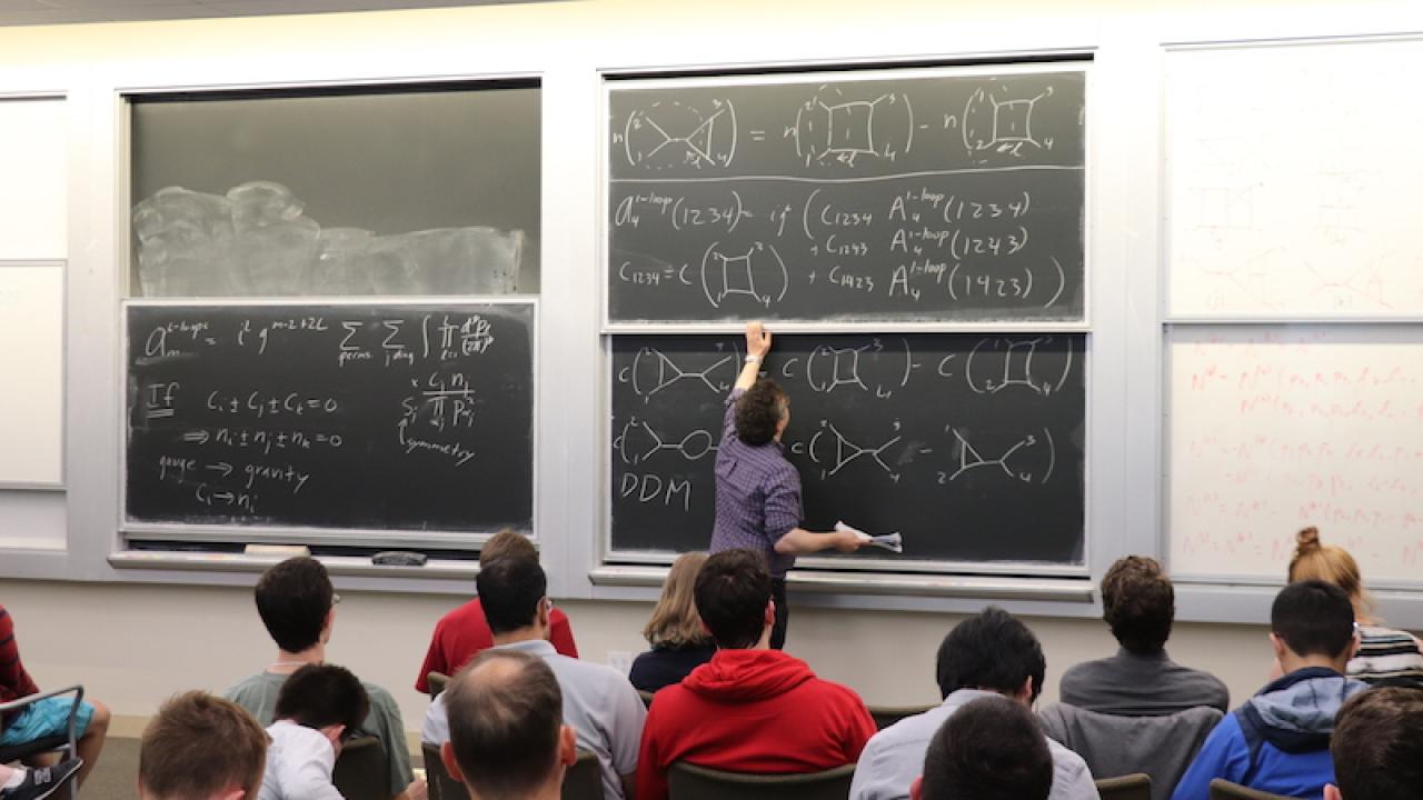 From the rear of a classroom, rows of students face a blackboard where a professor is drawing symbols. 