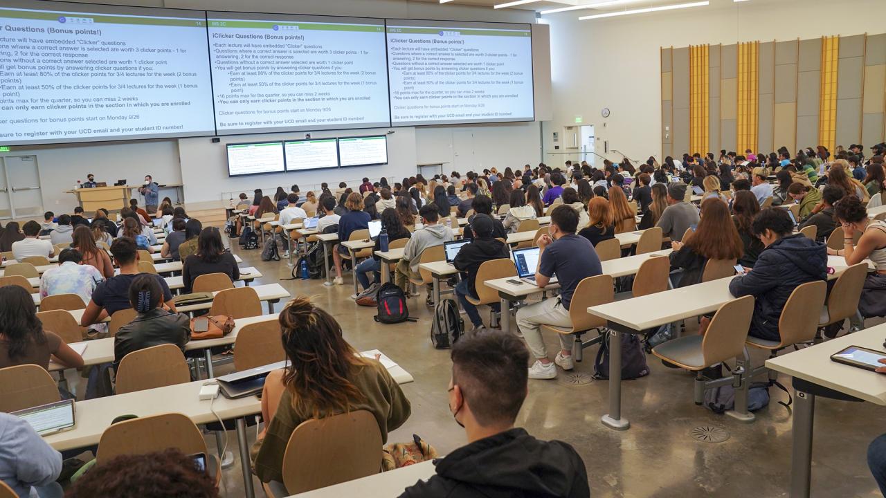 Seen from behind, students in tiered seats face three large screens at the front of a large lecture hall.