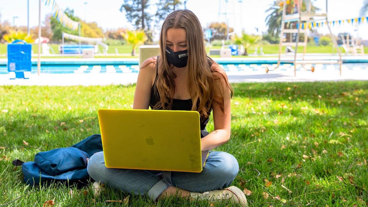 A student works on her laptop in a mask on the grass in front of a pool at UC Davis.