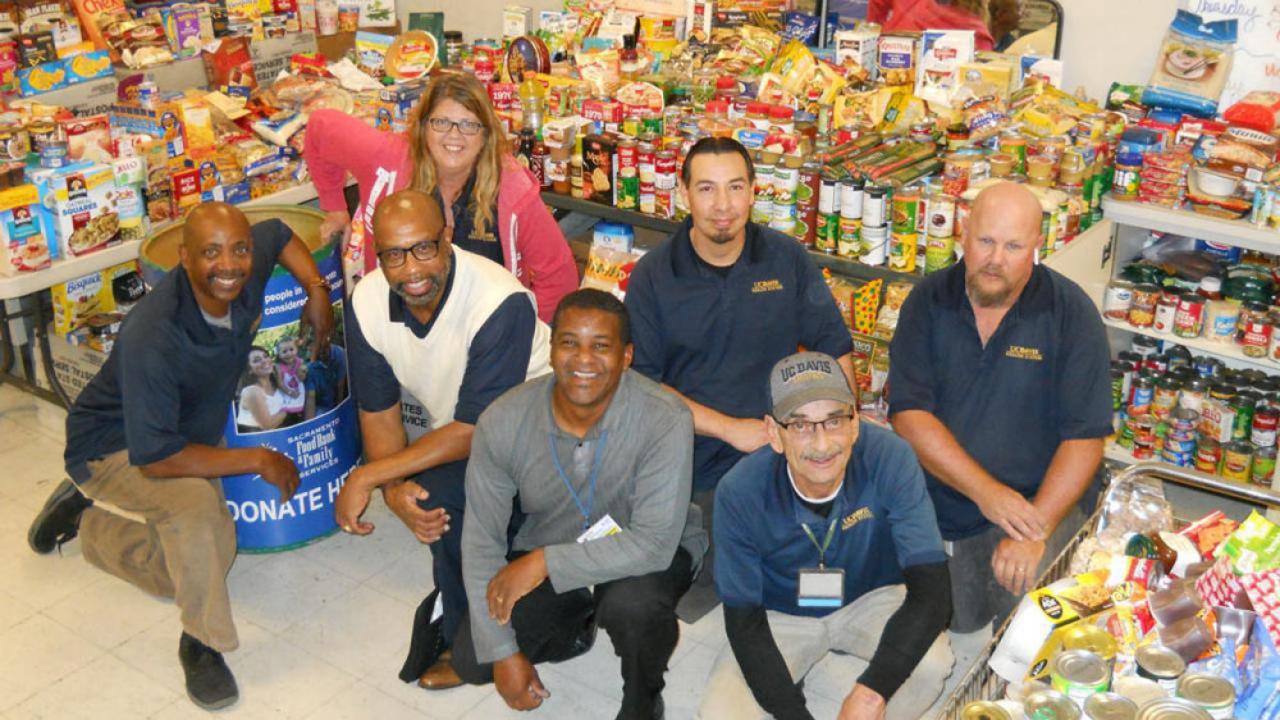 Seven employees pose in front of tables stacked with food donations.