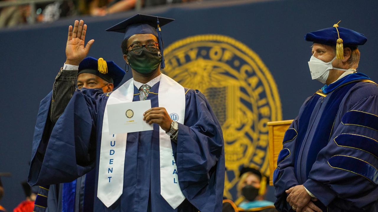Graduate in commencement regalia (and mask) waves as he walks across stage
