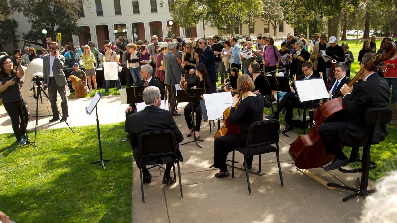 Members of a symphony play music near the Eye on Mrak Egghead sculpture as a large crowd looks on.