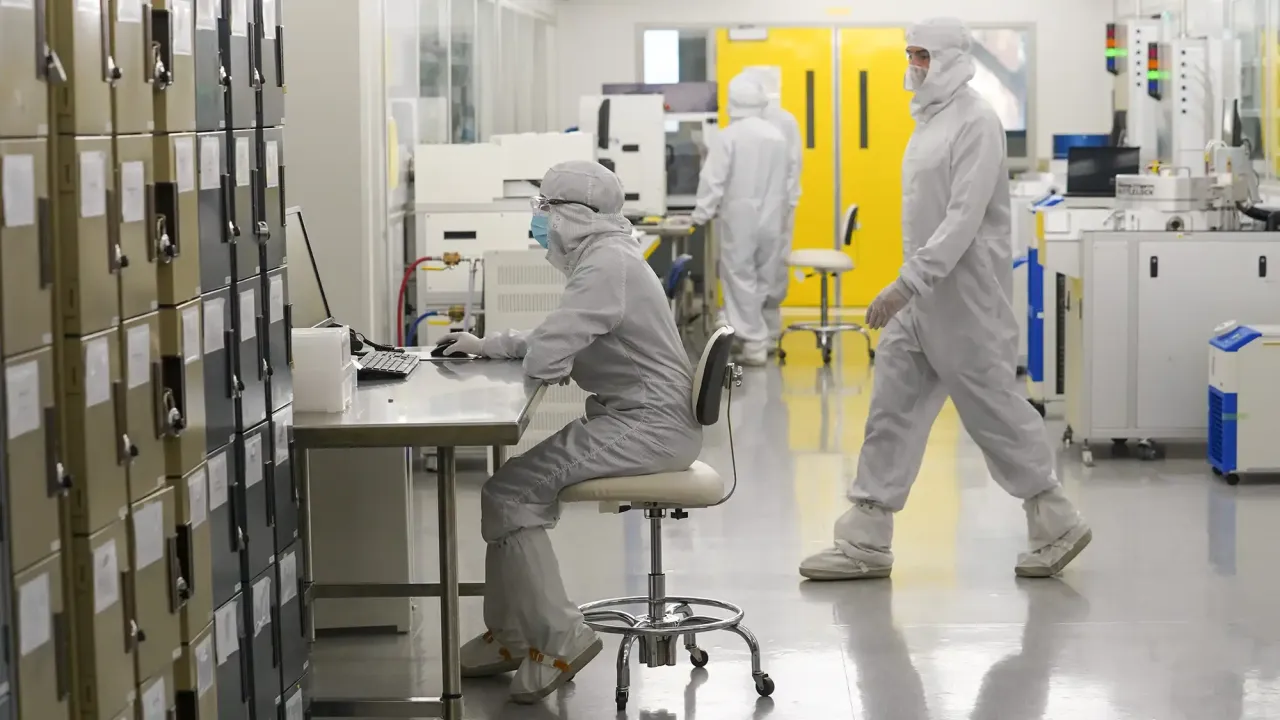 Researchers wearing full cleanroom suits work inside a bright, sterile laboratory. One person sits at a metal workstation using a computer, while others walk or operate equipment in the background. Rows of storage lockers line one side of the room, and large yellow doors are visible at the back.