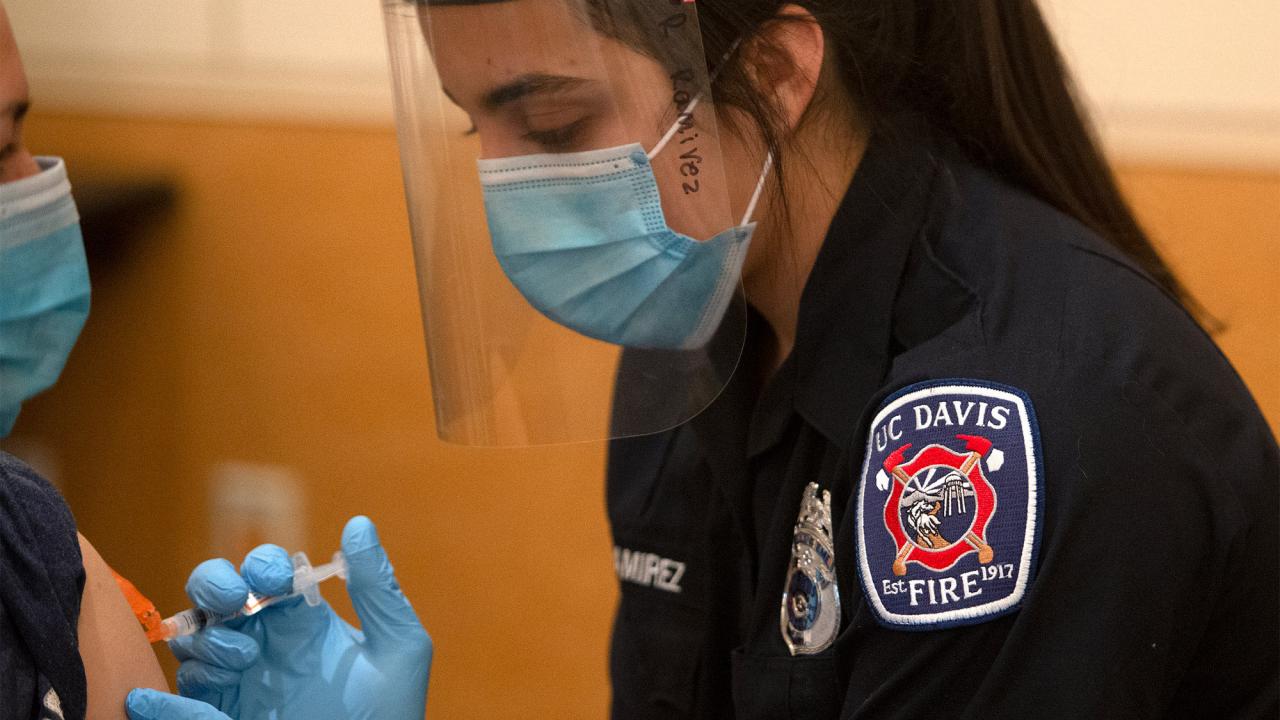 Student firefighter, in uniform, administers a vaccination.