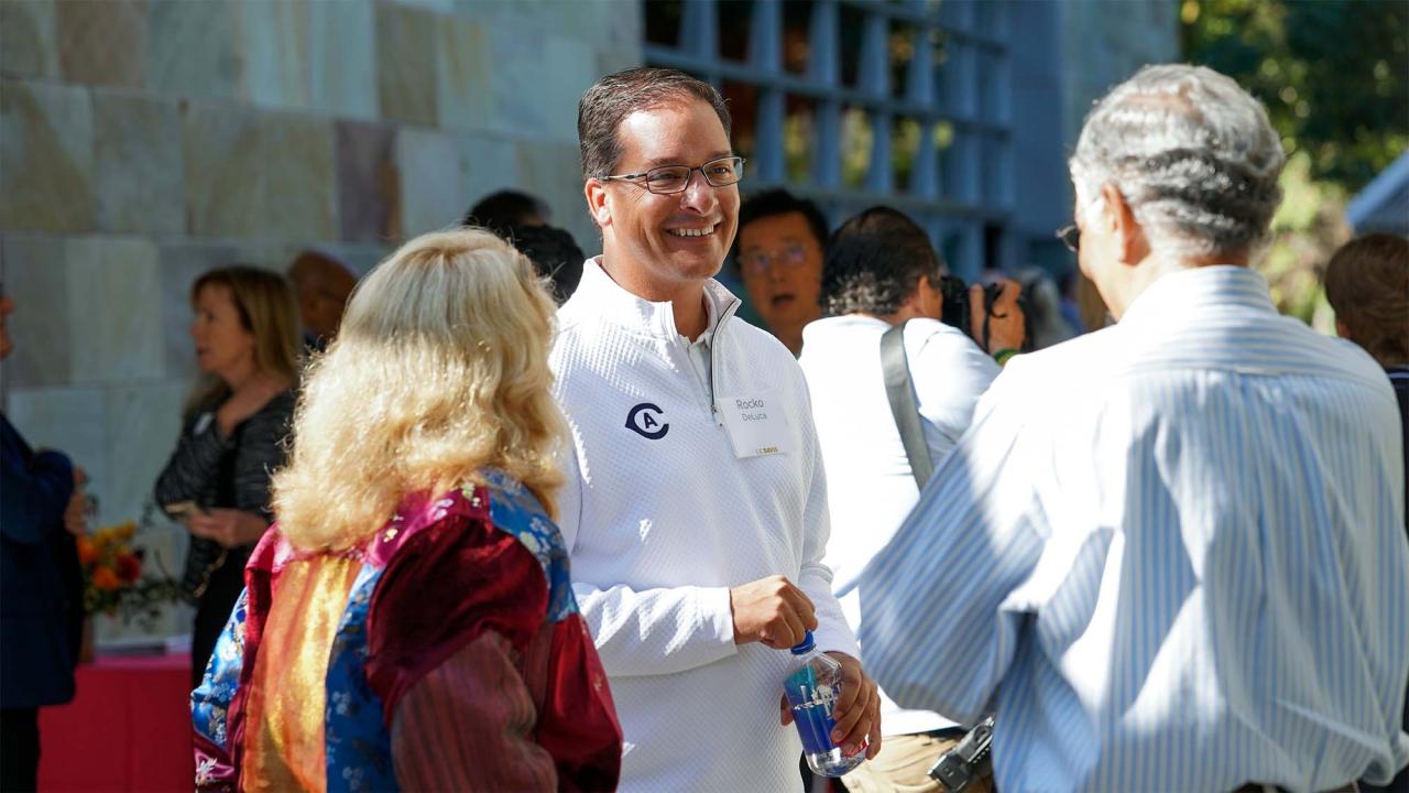 A smiling man in a white shirt converses with two people outdoors at a gathering.