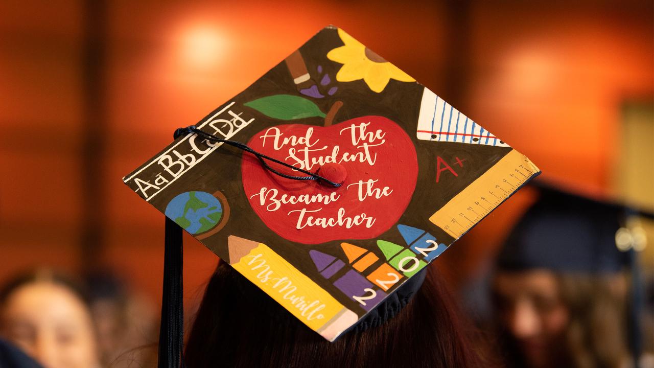 A grad cap that reads, "And the student became the teacher," in white cursive within a bright red apple, surrounded by teaching equipment like rulers, crayons and a pencil that reads, "Ms. Murillo." The photo was taken at UC Davis' School of Education Commencement graduation ceremony in 2023.