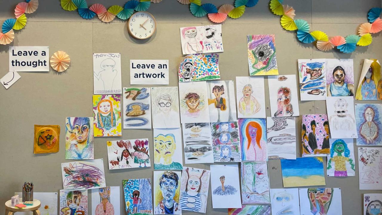 A wall in the ArtSpark studio is filled with drawings made by past visitors. The drawings include self-portraits, landscapes, and other abstract shapes.