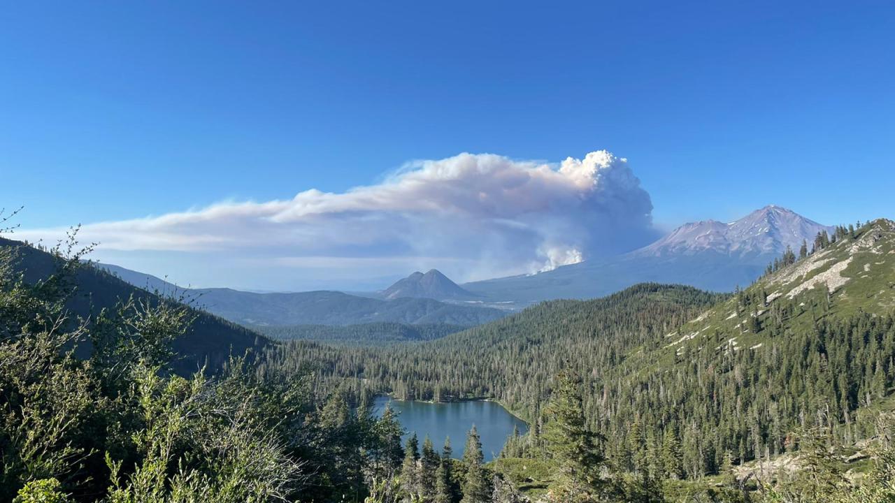 dramatic smoke plume billows over forested Castle Lake and Sierra Nevada mountains against blue sky