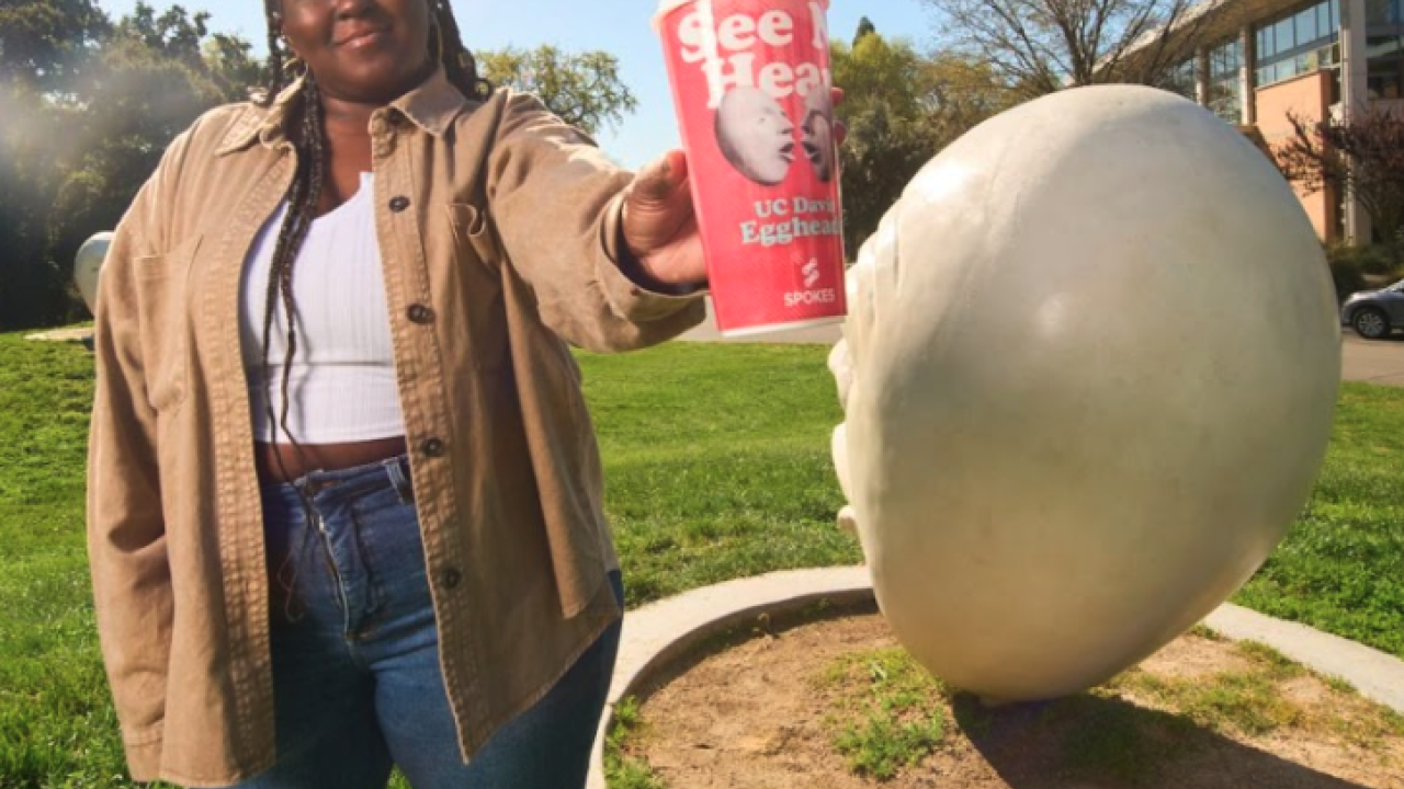 Aggie student stands in front of an Egghead sculpture while holding an Egghead Collectible Cup available at Spokes for a limited time