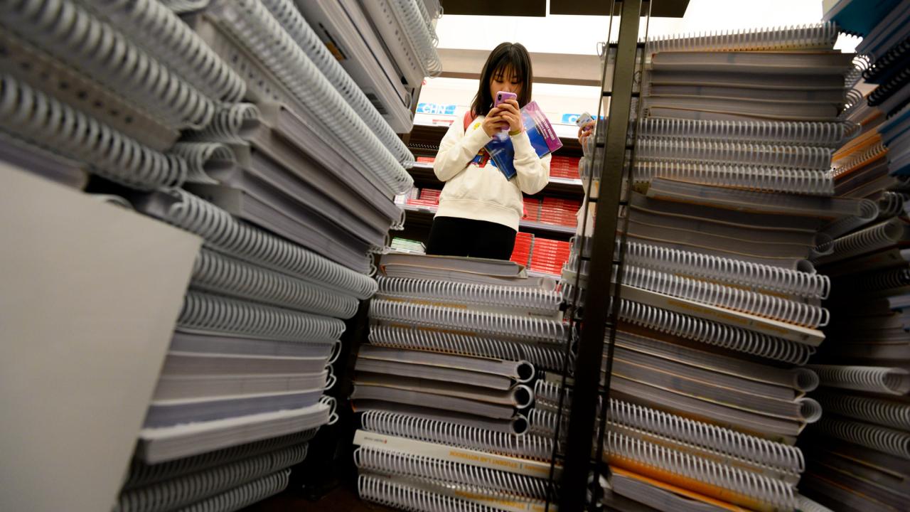 Person seen through stacks of notebooks, looking at phone