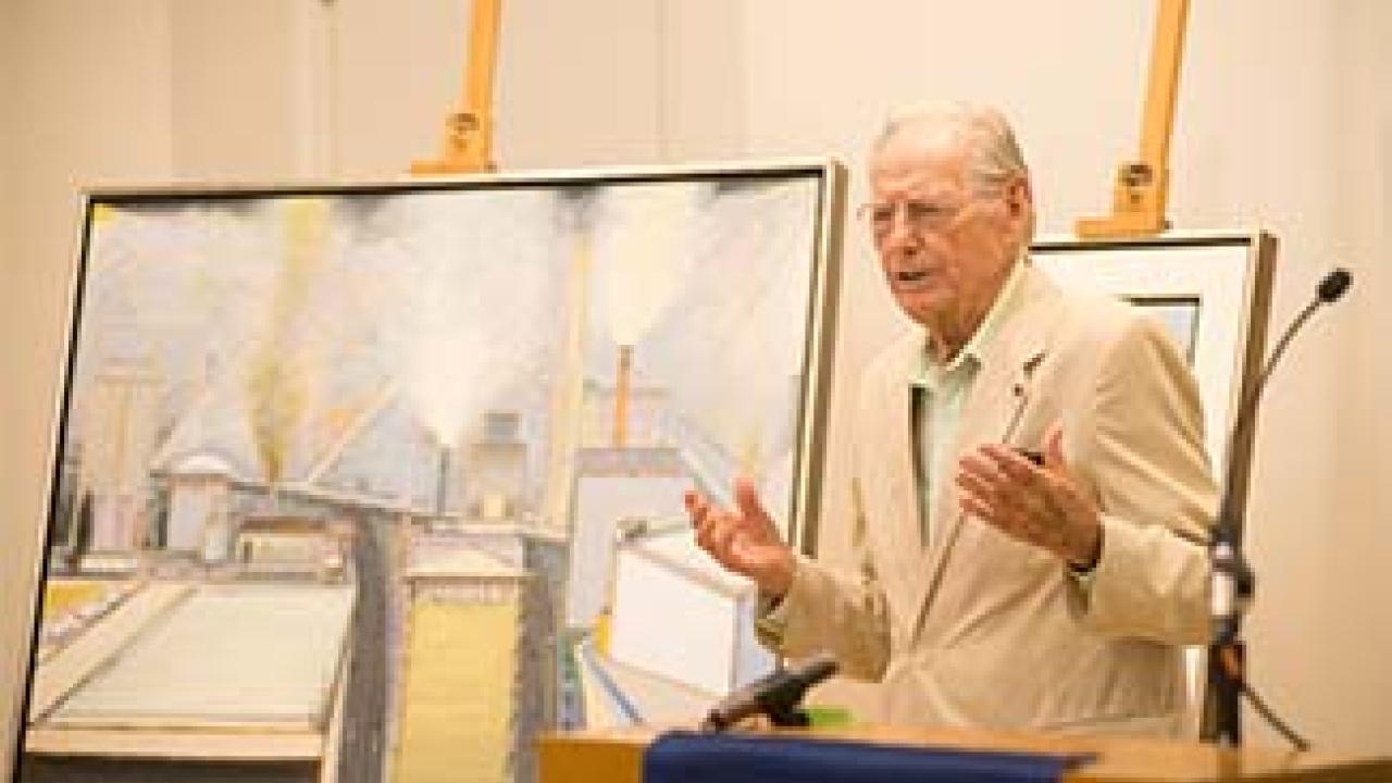 Man stands in front of paintings, addresses audience.