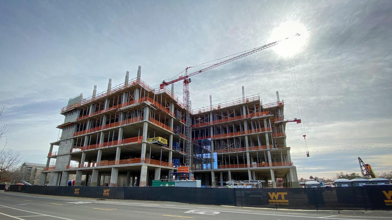 Aggie Square, a UC Davis building on the Sacramento campus, is undergoing construction. The building's framing comprised of structural beams peaks into the partially cloudy sky.